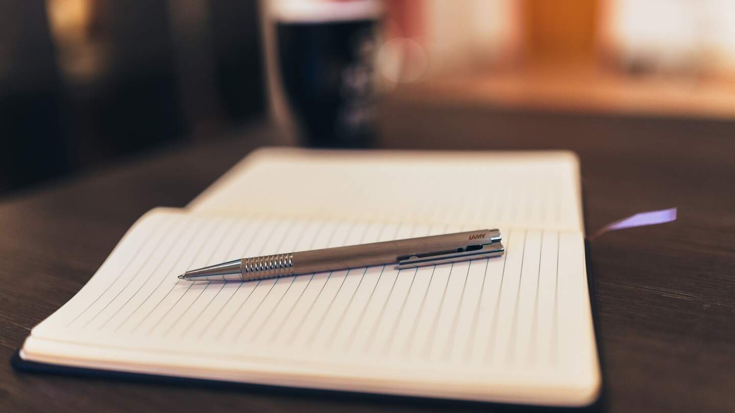 Ein geoeffnetes, unbeschriebenes Notizbuch und ein Kugelschreiber liegen im Vordergrund auf einem braunem Holztisch, im Hintergrund steht eine blaue Tasse | © Pixabay.com/stocksnap