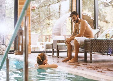 Mann und Frau unterhalten sich im Hallenbad. Die Frau ist in einem Pool und der Mann sitzt auf einem Liegestuhl. | © Gettyimages.com/fotostorm
