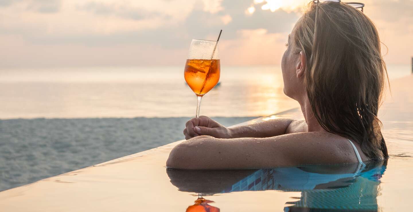 Frau trinkt Cocktail bei Sonnenuntergang in einem Infinity-Pool | © © Gettyimages.com/Mystockimages