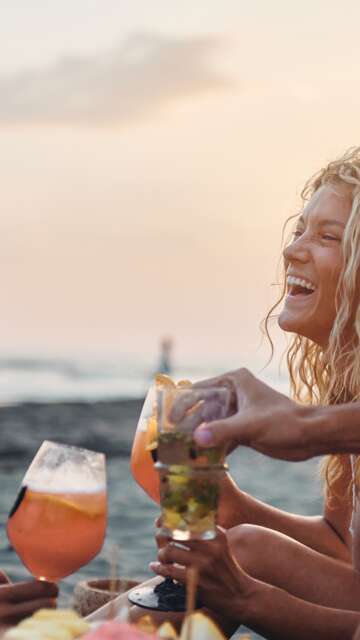  Fröhliche Freunde genießen Obst und Getränke am Strand bei Sonnenuntergang im Sommerurlaub. | © GettyImages.de/skynesher