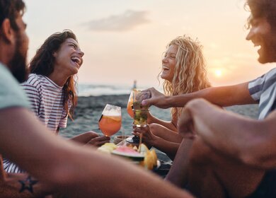  Fröhliche Freunde genießen Obst und Getränke am Strand bei Sonnenuntergang im Sommerurlaub. | © GettyImages.de/skynesher
