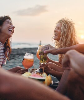  Fröhliche Freunde genießen Obst und Getränke am Strand bei Sonnenuntergang im Sommerurlaub. | © GettyImages.de/skynesher