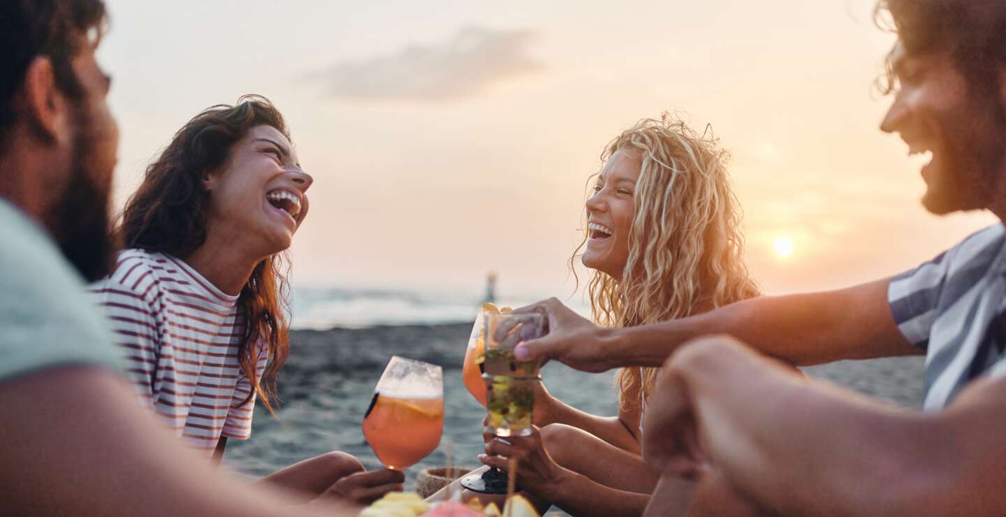  Fröhliche Freunde genießen Obst und Getränke am Strand bei Sonnenuntergang im Sommerurlaub. | © GettyImages.de/skynesher