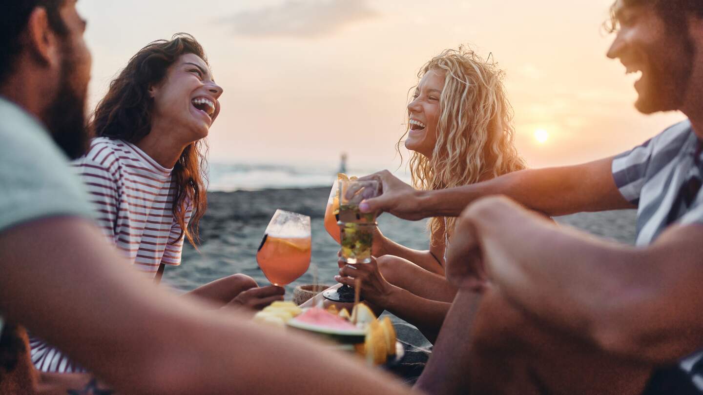  Fröhliche Freunde genießen Obst und Getränke am Strand bei Sonnenuntergang im Sommerurlaub. | © GettyImages.de/skynesher