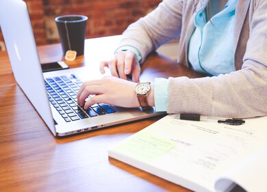 Angeschnittener Oberkoerper einer Frau, die auf ihrem Laptop tippt und ihren arm auf einem geoeffneten Heft mit Notizen liegt, neben ihr ihre Teetasse und ihr Handy | © Pixabay.com/StartupStockPhotos