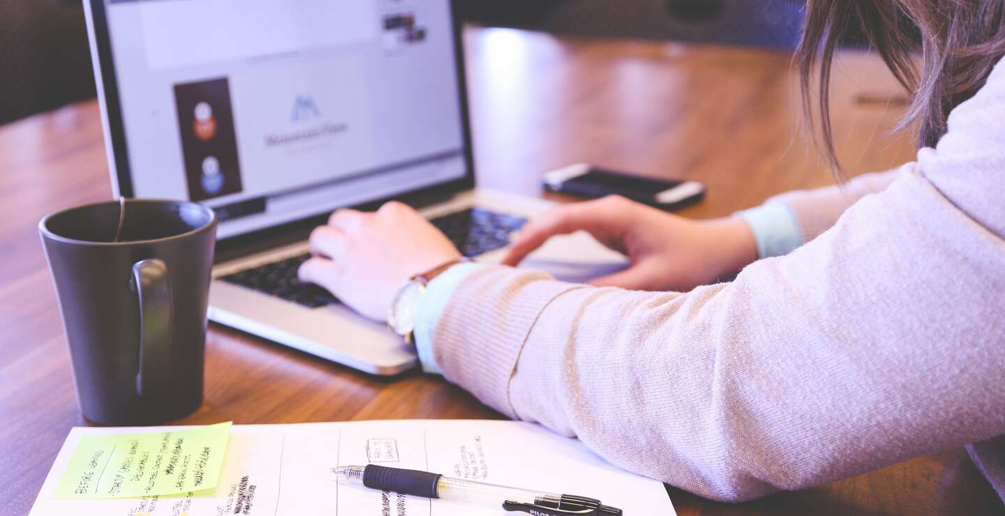 Angeschnittene Frau, die auf einem geoeffneten Laptop tippt, waehrend neben ihr liegt ein Blatt mit Notizen und eine Teetasse | © Pixabay/StartupStockPhotos 