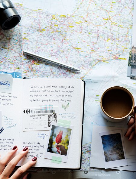 eine flache Anordnung verschiedener Reiseutensilien auf einer Oberflaeche, darunter eine Karte, ein Fernglas, ein Reisepass, Postkarten, Fotos und eine Tasse Kaffee | © gettyimages.com/Rawpixel Ltd.