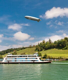 Passagierfähre von Meersburg auf ihrer Route über den Bodensee an einem Sommertsg, mit Weinbergen und einem Zeppelin im Hintergrund | © © Gettyimages.com/olgysha2008