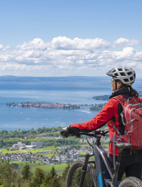 Frau mit Elektro-Mountainbike steht auf einem Berg und schaut hinunter auf das Panorama des Bodensees | © GettyImages.com/Uwe Moser