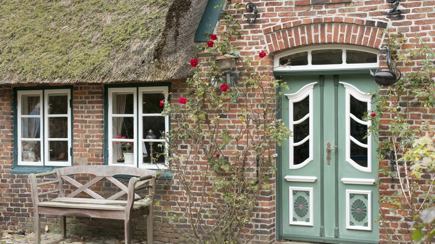 Fassade eines typischen traditionellen Zuhauses auf der Insel Sylt, Deutschland. Die grüne Tür wird von roten Rosen umramt. Vor dem Haus steht eine BAnk | © Gettyimages.com/relaxfoto.de