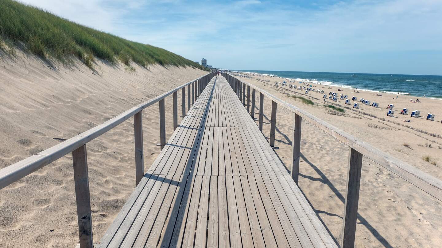 Die bekannte Strandpromenade mit dem Holzsteg in Richtung der Stadt Westerland, Hauptstadt der deutschen Insel Sylt in der Nordsee. Rechts der Strand voll mit Sttr4andkörben und Menschen, links die Düne | © Gettyimages.com/karlhendriktittel