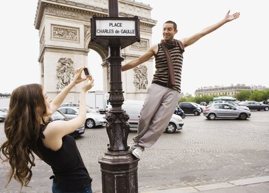 Frau nimmt vor dem Arc de Triomphe ein Foto von einem Mann auf, der auf einem Straßenschild steht.  | © Gettyimages.com/Jupiterimages
