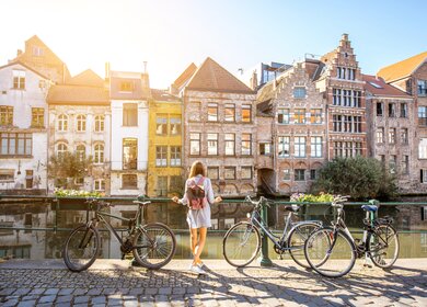 Frau in Gent, Belgien mit Fahrrädern | © Gettyimages.com/RossHelen Eine Frau steht in Gent, Belgien an einem Fluss und blickt auf die Altstadt. Neben ihr stehen Fahrräder | © Gettyimages.com/RossHelen
