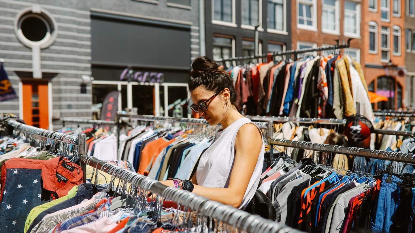 Shoppen auf einem Flohmarkt in Amsterdam | © Gettyimages.com/lechatnoir Frau mit Sonnenbrille und ärmelloser Bluse schaut durch die Kleiderstaender eines Flohmarktes in Amsterdam | © Gettyimages.com/lechatnoir