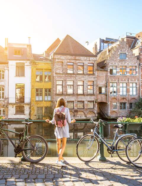 Eine Frau steht in Gent, Belgien an einem Fluss und blickt auf die Altstadt. Neben ihr stehen Fahrräder | © © Gettyimages.com/RossHelen