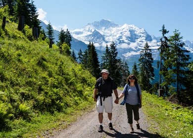 Wandern im berner Oberland | © Gettyimages.com/OGphoto Reifes Paar, das Haendchen haelt, wandert einen Bergpfad hinunter mit schneebedeckten Bergen hinter sich. Im Berner Oberland bei Wengen, Schweiz. | © Gettyimages.com/OGphoto