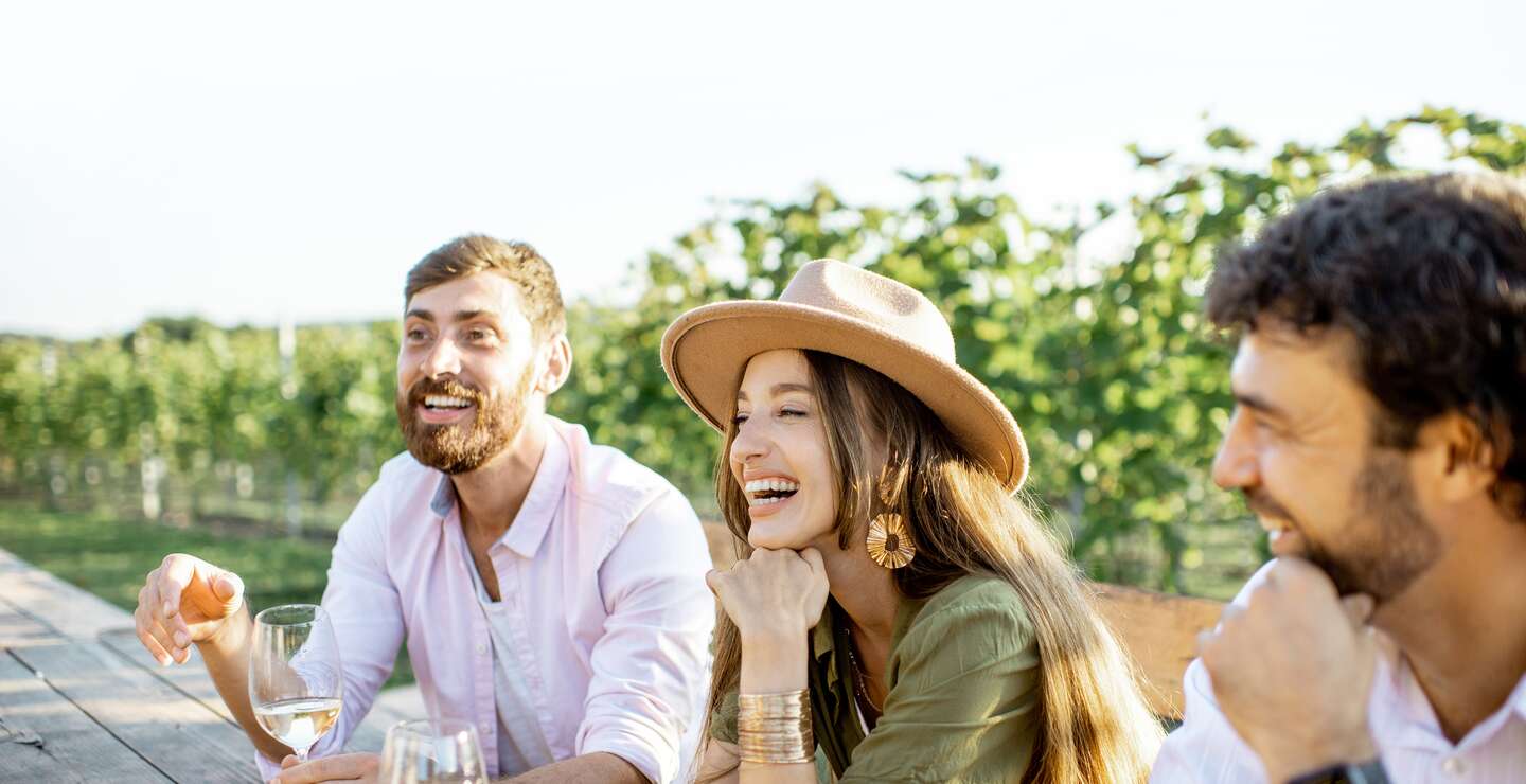 Menschen, die Wein trinken und sich miteinander unterhalten, während sie an einem sonnigen Abend am Esstisch im Freien auf dem Weinberg sitzen | © Gettyimages.com/RossHelen