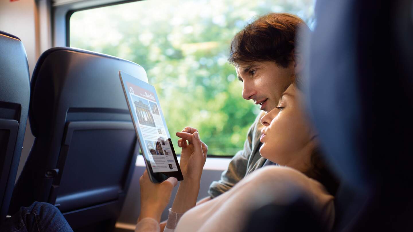 Paar im ICE | ©  Deutsche Bahn AG  eine Person, die in einem Zug sitzt und auf ein Tablet schaut, auf dem Text und Bilder zu sehen sind.  | ©  Deutsche Bahn AG