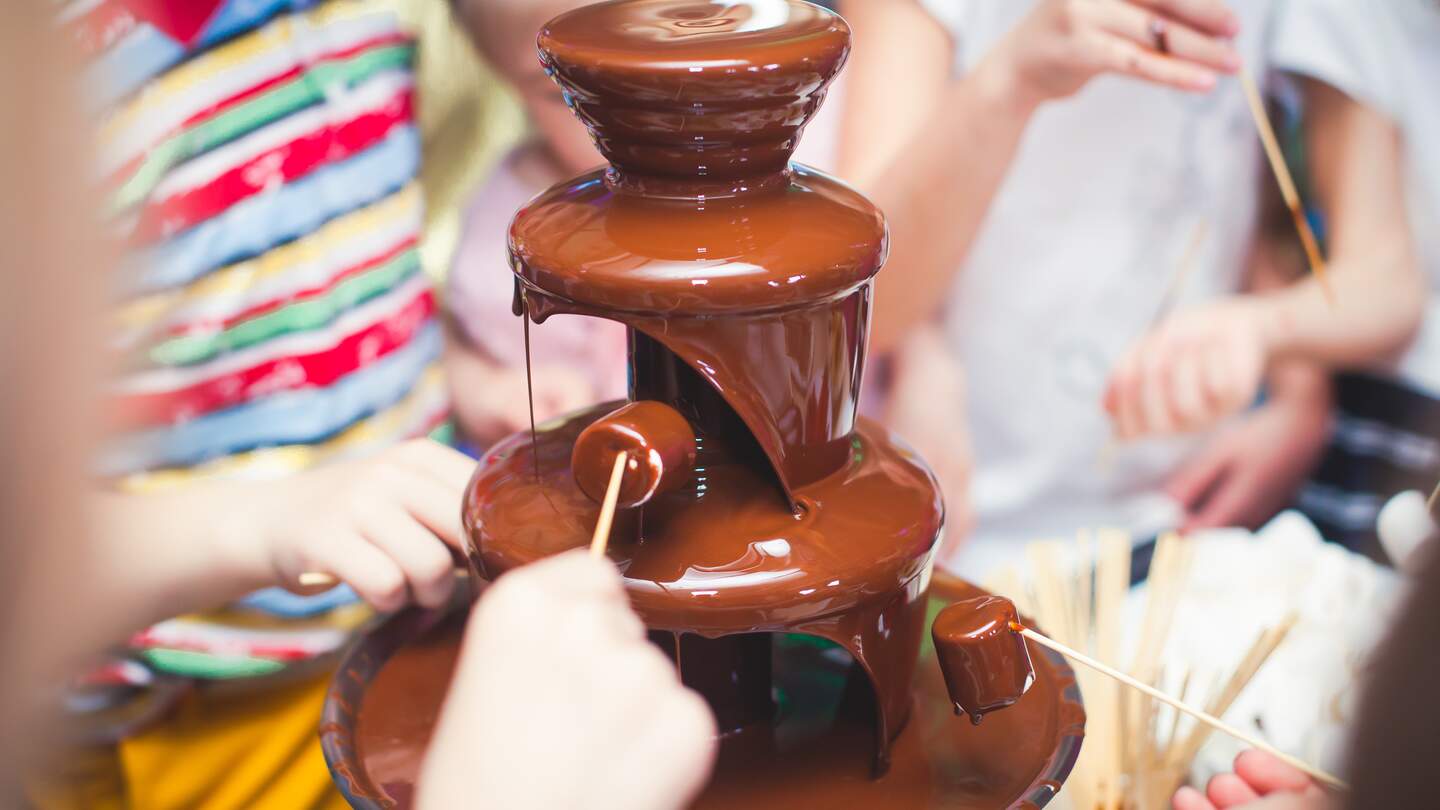 Lebendiges Bild von Chocolate Fountain Fontain auf einer Kindergeburtstagsparty mit herumspielenden Kindern und Marshmallows und Früchten, die in den Brunnen tauchen | © Gettyimages.com/tsguliev