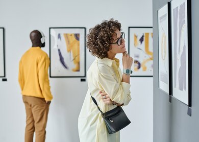 Moderne Kunst in der Galerie  | © Gettyimages.com/AnnaStills Junge Frau mit Brille untersucht moderne Kunst in der Galerie mit anderen Besuchern | © Gettyimages.com/AnnaStills