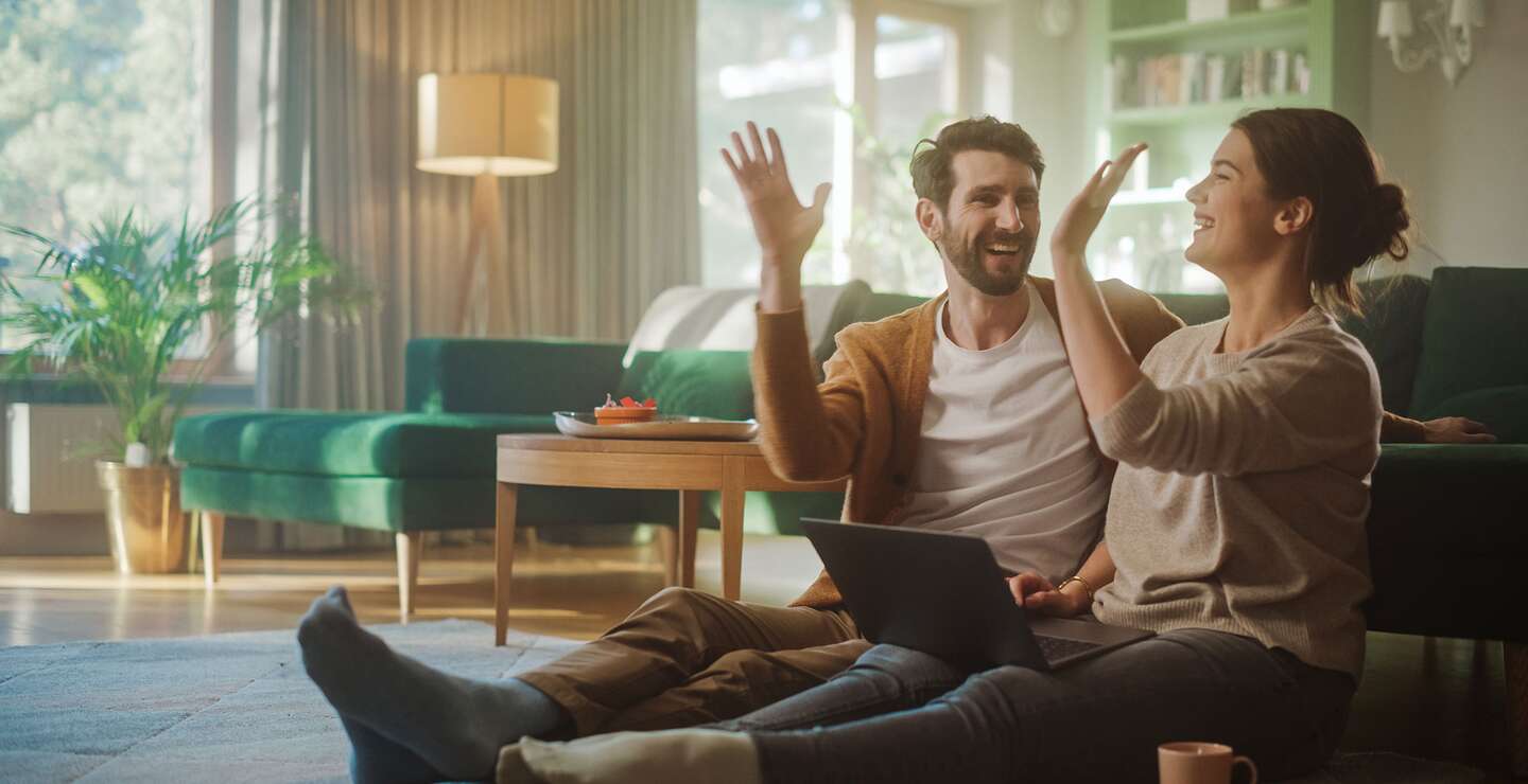 Paar gibt sich High Five | ©  Gettyimages.com/gorodenkoff Paar benutzt Laptopund gibt sich ein High-Five, während sie im Wohnzimmer auf dem Fussboden sitzen | ©  Gettyimages.com/gorodenkoff