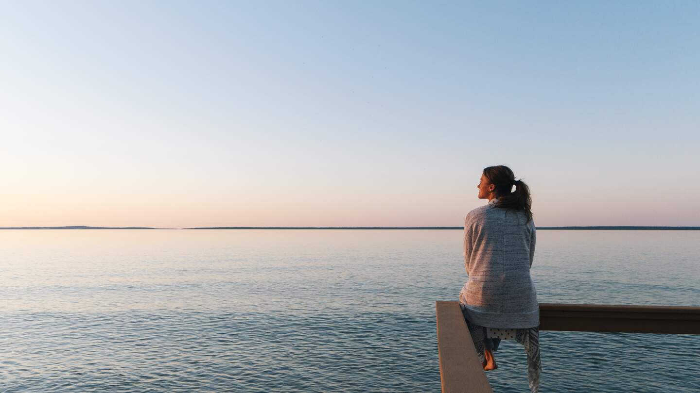 Nachdenkliche Frau sitzt am Wasser und blickt in die Ferne und genießt die ruhige Atmosphäre | © Gettyimages.com/AscentXmedia