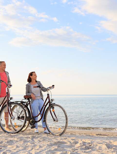 Ein Paar steht mit Fahrrädern am Sandstrand einer Küste mit blauem Wasser und hellem Himmel | © GettyImages.com/FredFroese