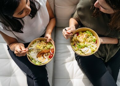 Zwei Kolleginnen sitzen auf einem hellen Sofa und essen eine gesunde und ausgewogene Bowl mit Salat zu Mittag | © Gettyimages.com/AnVr