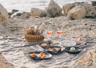 Strandpicknik | © gettyimages.com/Polina Lebed ein Picknick-Setup an einem Sandstrand mit Felsen im Hintergrund,darunter  ein Weidenkorb, Teller mit Essen, Besteck, Weinglaese | © gettyimages.com/Polina Lebed